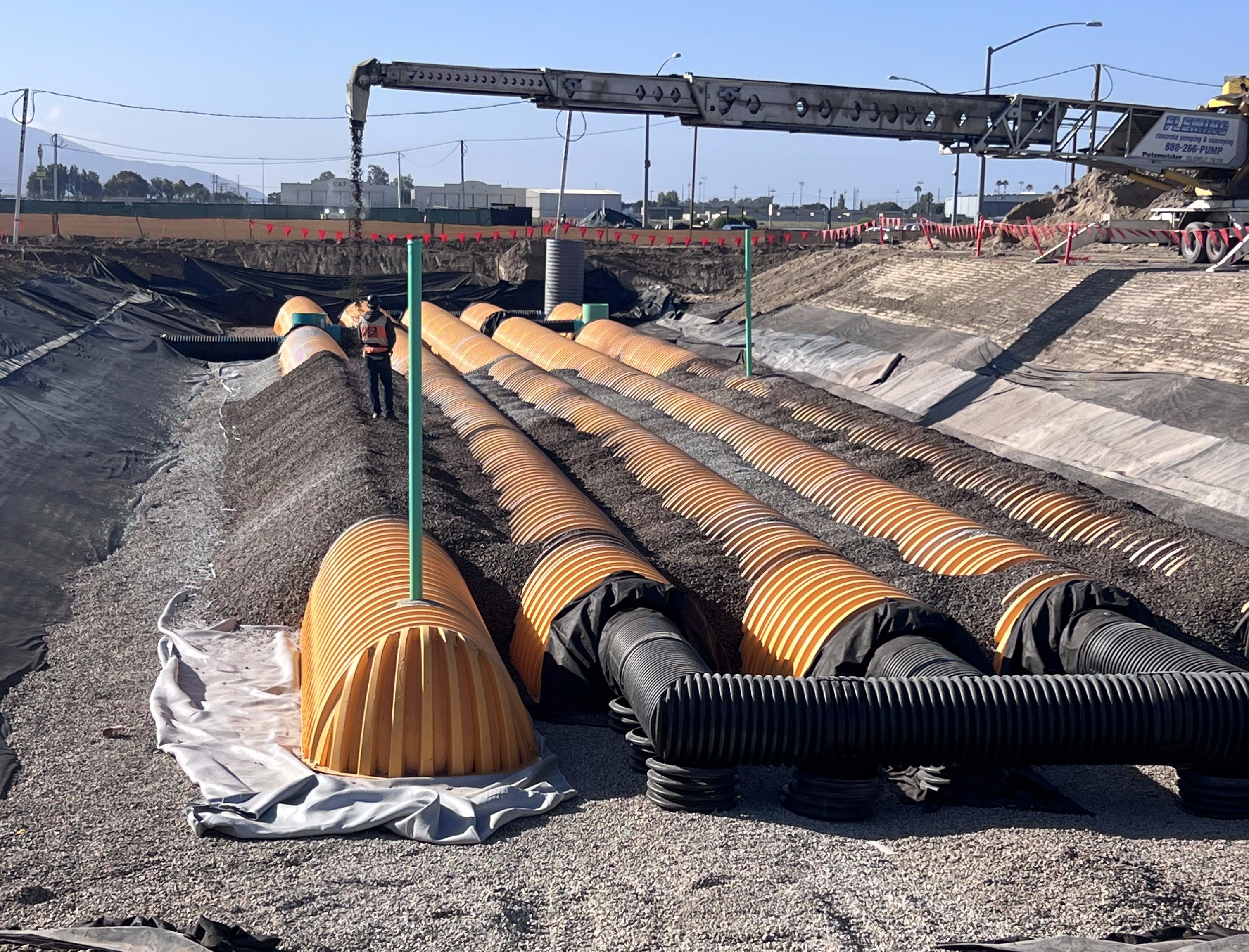 Installation of an underground stormwater detention system featuring rows of orange corrugated chambers surrounded by stone aggregate at a Costco warehouse construction site. A worker stands between the chambers as equipment places additional stone.