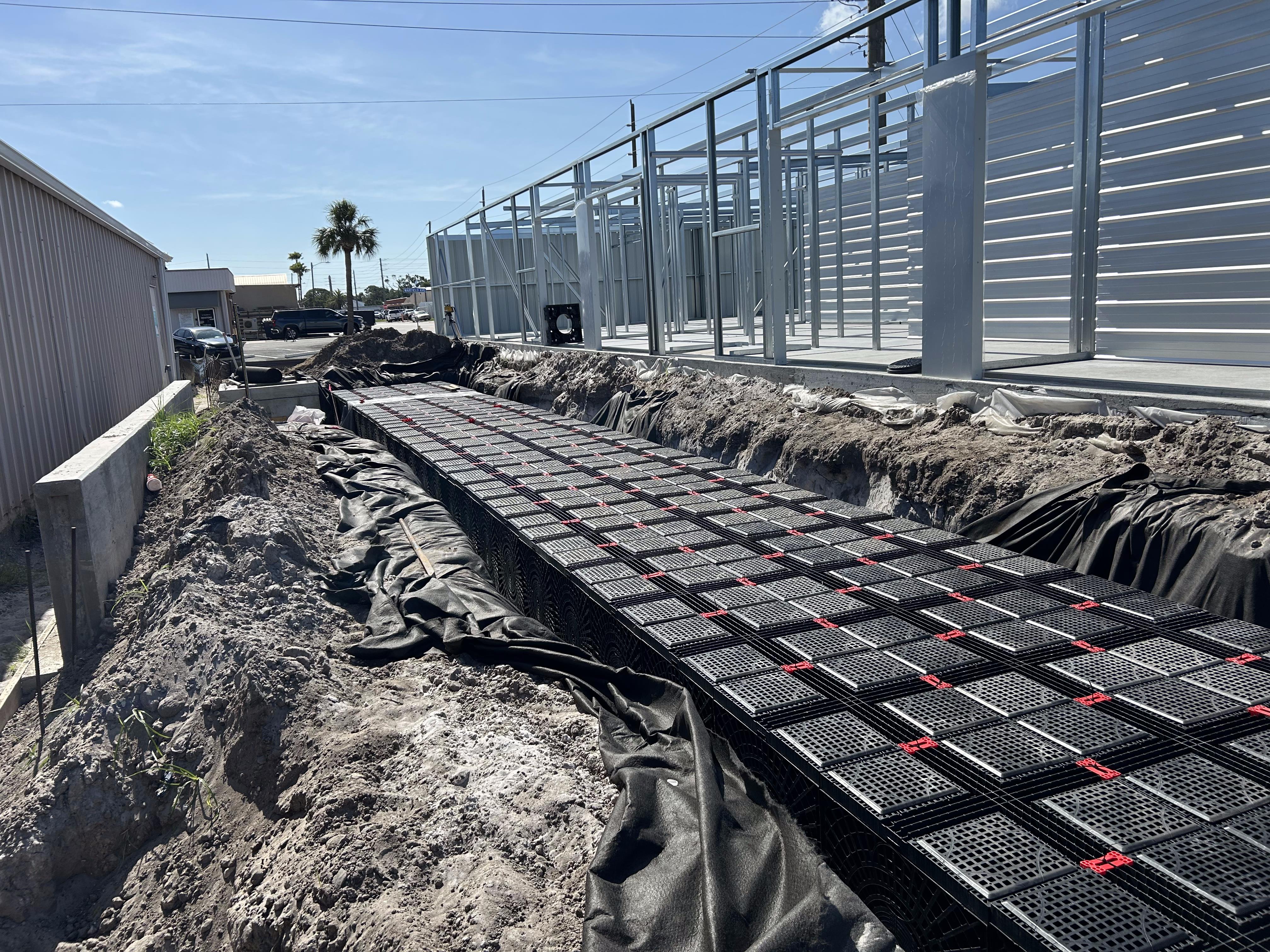 Underground stormwater detention system installed in a trench beside a metal building under construction, showing modular plastic chambers wrapped in geotextile fabric before backfill.
