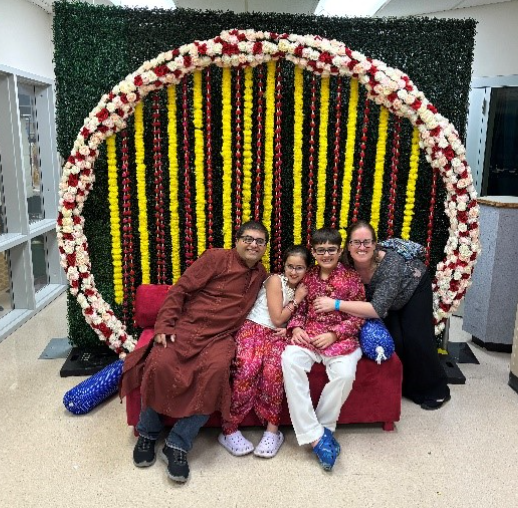 A family of four poses together on a red couch in front of a vibrant floral backdrop made of yellow, red, and white garlands arranged in a circular arch. 
