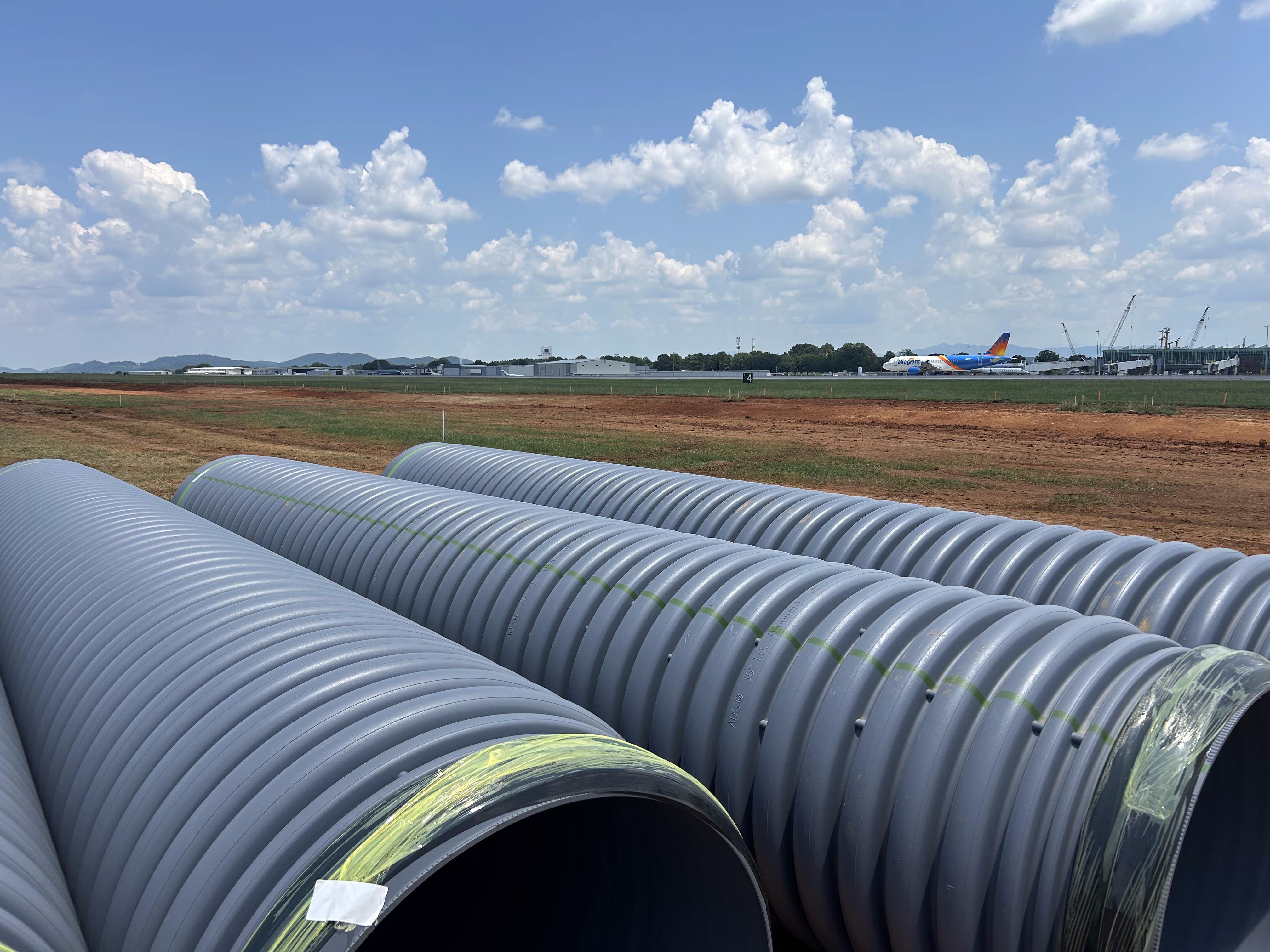 Large gray corrugated drainage pipes are stacked on a construction site near an airport runway.