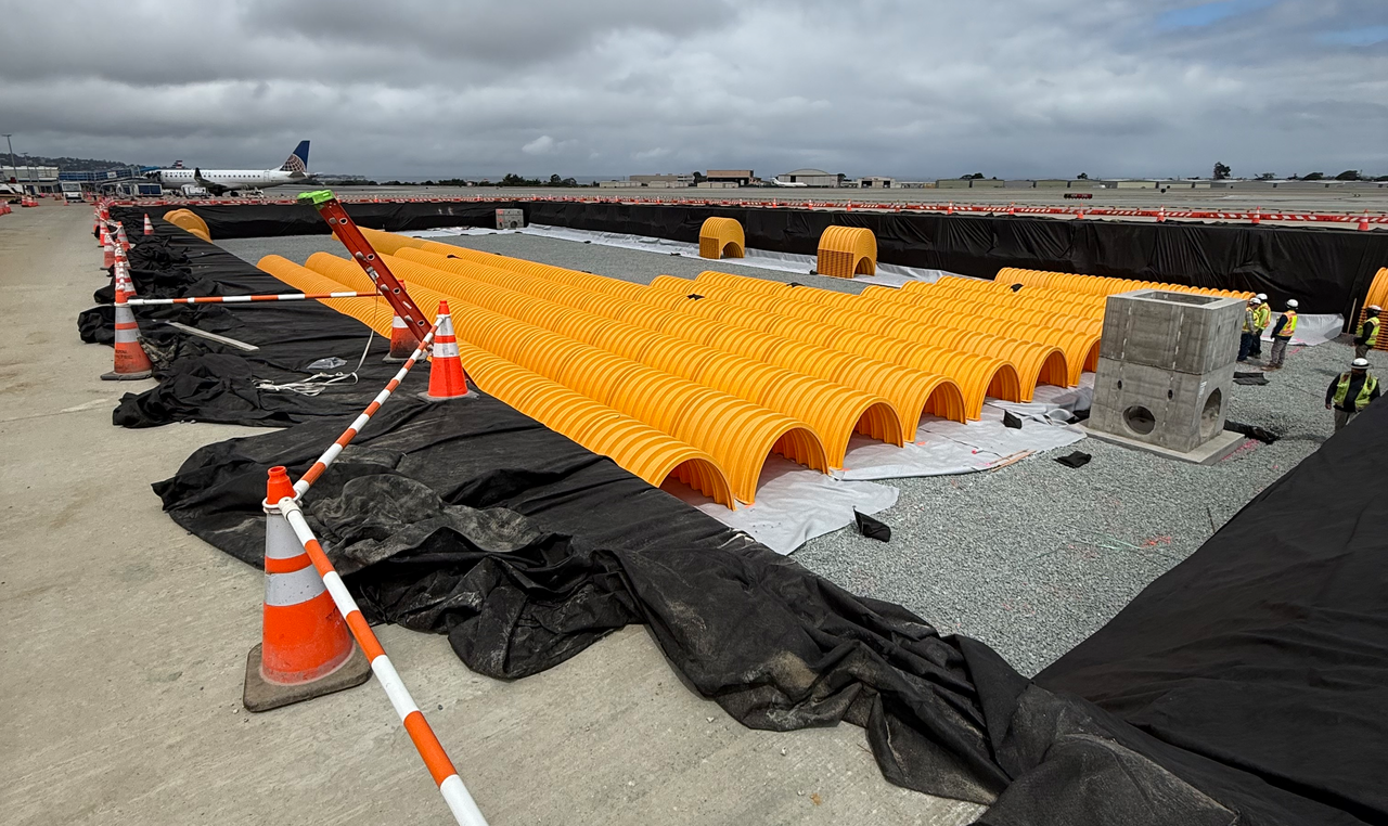 A stormwater chamber system installation at an airport construction site. Rows of bright yellow plastic chambers are laid out on a gravel bed, surrounded by black geotextile fabric.