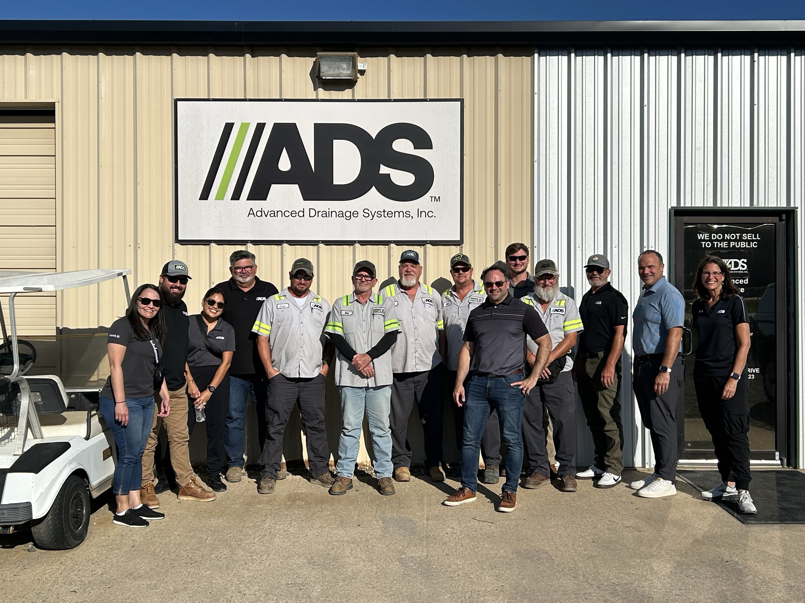 Group of ADS employees and drivers standing together outside an Advanced Drainage Systems facility, smiling in front of a large ADS sign.