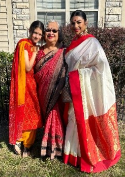 Three women stand together outside in front of a house, smiling warmly. They are dressed in traditional South Asian attire. 
