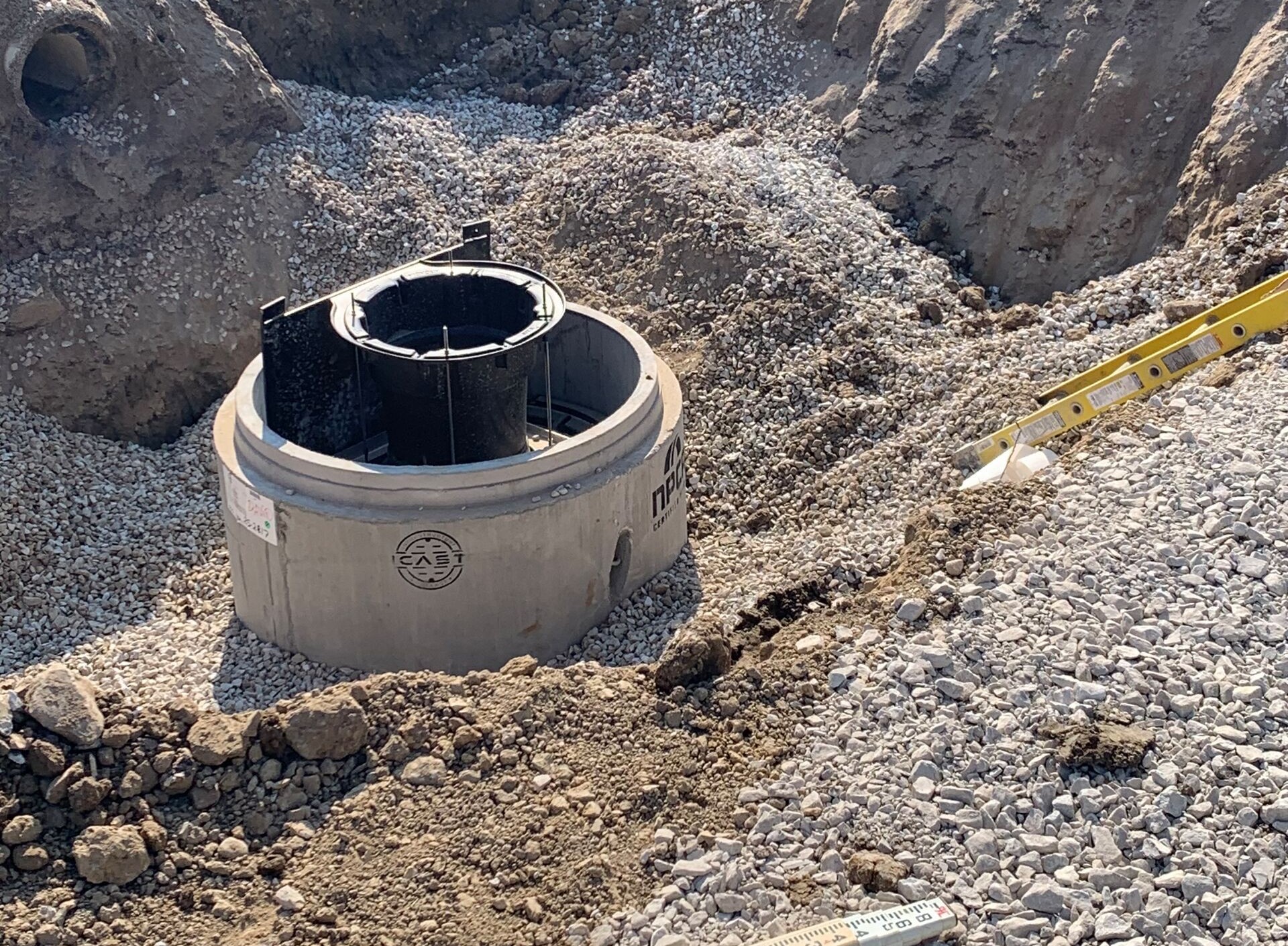 Precast concrete manhole installed in an excavated trench, surrounded by crushed stone bedding, with a black internal insert visible and a level resting on the gravel during installation.